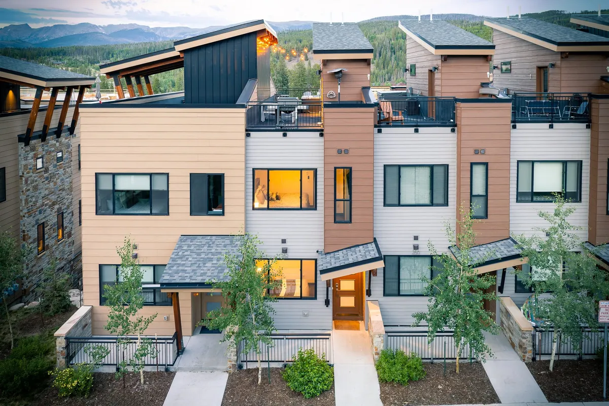 The image showcases the striking modern architecture of a luxury single-family villa, highlighted by a spacious balcony with inviting seating, lush landscaped greenery, and the promise of colorful sunset views from the rooftop. The scene radiates warmth and sociability, suggesting the perfect spot for outdoor dining, relaxing with friends and family, and embracing mountain adventures just beyond the doorstep.