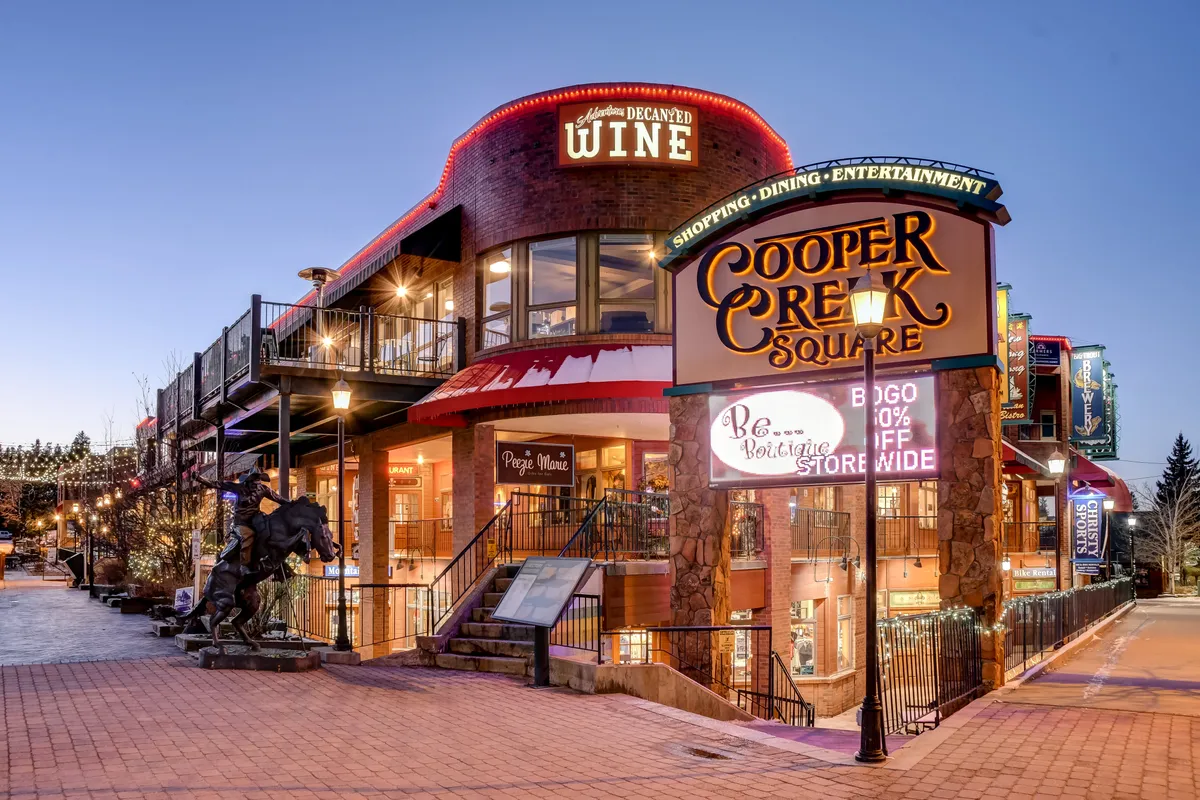 This vibrant exterior image shows the Cooper Creek Square sign, a wine store entrance, an outdoor seating area, and decorative lighting. People can be seen shopping, dining, and socializing, contributing to a lively, festive atmosphere that captures the essence of downtown Winter Park's social scene.