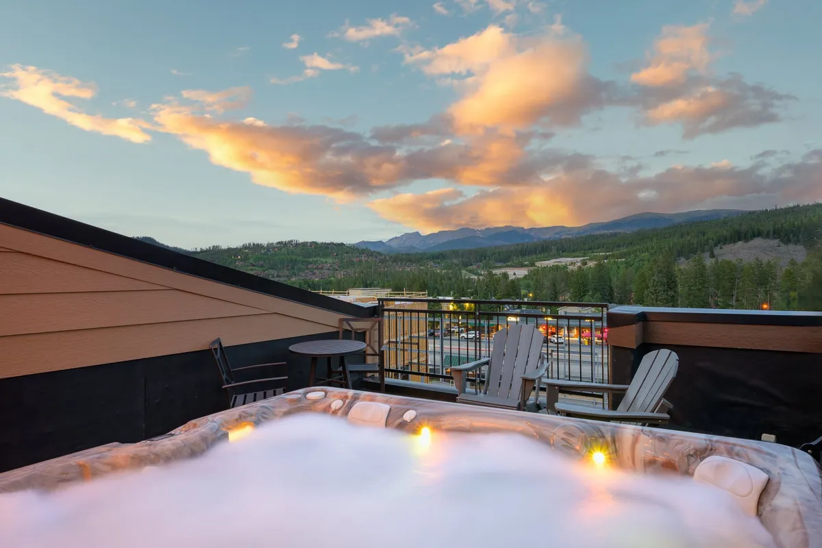 This exterior image captures a private rooftop hot tub with panoramic mountain views, surrounded by outdoor seating. The tranquil setting is highlighted by clouds drifting above the peaks, inviting relaxation and socializing. The arrangement of seating suggests space for both intimate gatherings and group enjoyment.