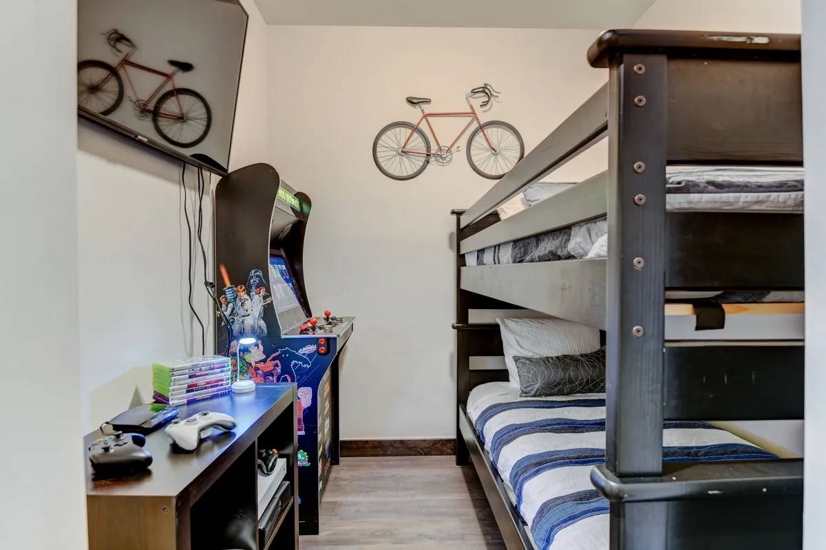 A playful, youth-oriented living space featuring bunk beds adorned with colorful bedding, wall-mounted decorative bicycles, a custom arcade machine, and a gaming console area. The environment is lively and designed for both sleepovers and active play, with plenty of room for family bonding activities.