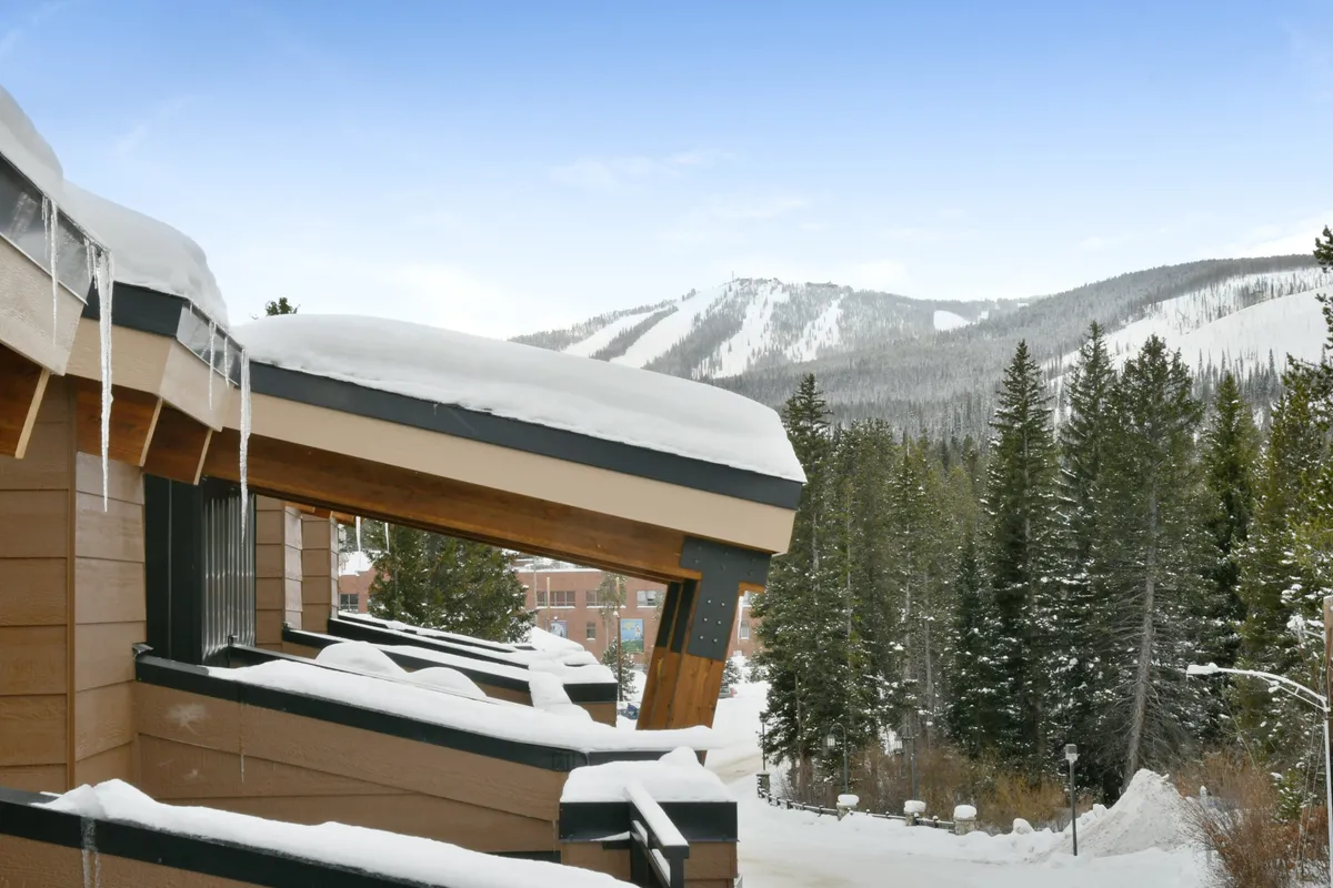 A picturesque winter exterior with snow-covered roofs surrounded by pine trees, set against distant mountain peaks and a clear blue sky. The scene captures skiers and snowboarders enjoying the slopes, as well as relaxed moments like building snowmen and gathering around a fireplace after a day outdoors.