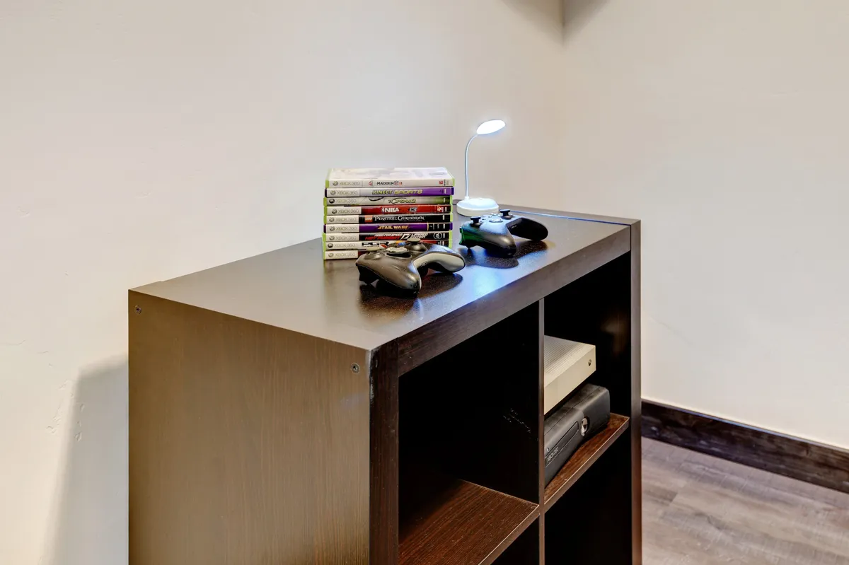 A cozy nook featuring a shelving unit stocked with various gaming consoles, a stack of video games, a table lamp, and game controllers. The setup invites casual gaming sessions, socializing, and relaxed family time.