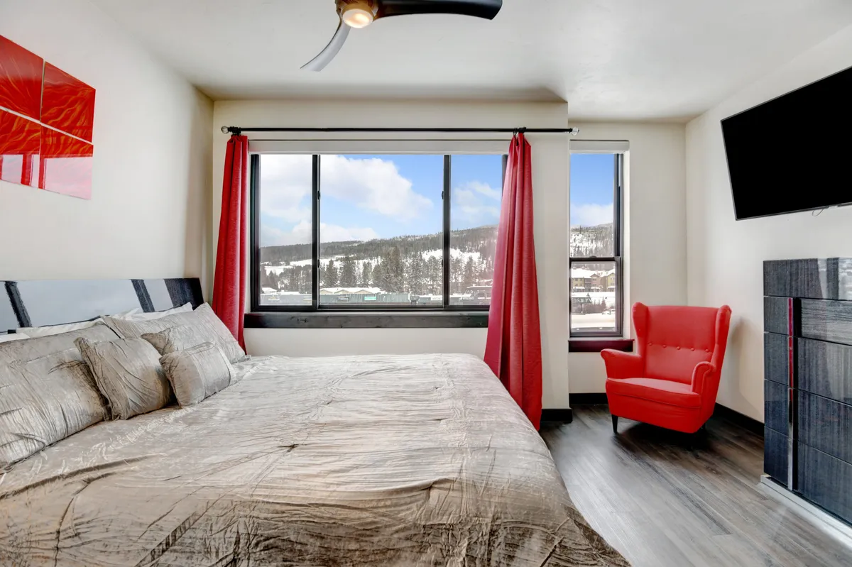 A tranquil bedroom with a comfortable bed, striking red accent chair, and large windows that let in natural light and offer mountain views. The setting is peaceful and ideal for unwinding with a book or enjoying the view.