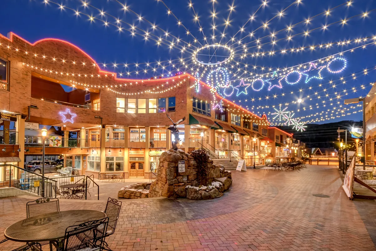 A charming downtown plaza is shown adorned with string lights and decorative stars. A central fountain, brick pathways, and storefronts frame the scene, with people shopping, dining, and enjoying the festive atmosphere. The mood is joyful and community-focused, evoking seasonal markets and local events.