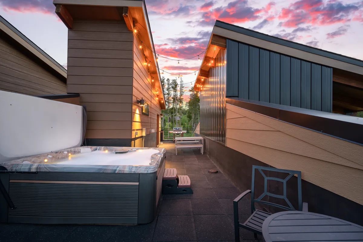 A rooftop setting featuring a bubbling hot tub with inviting steam rising, surrounded by cozy outdoor seating. Overhead, string lights cast a warm glow, and there's a clear view of the surrounding mountains in the background, suggesting both privacy and panoramic vistas.