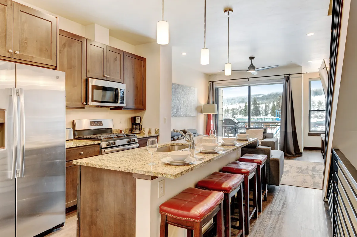 The image features a cozy, welcoming kitchen space with a granite countertop, bar stools, and a large window offering scenic views. The breakfast bar setup suggests a place for morning coffee or casual meals, and the warm lighting creates an inviting atmosphere.