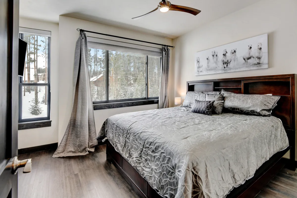 A spacious king bedroom featuring a large bed with premium bedding, expansive windows overlooking serene forest views, and tasteful artwork above the headboard. The room is bathed in natural light, creating a relaxing and inviting ambiance.