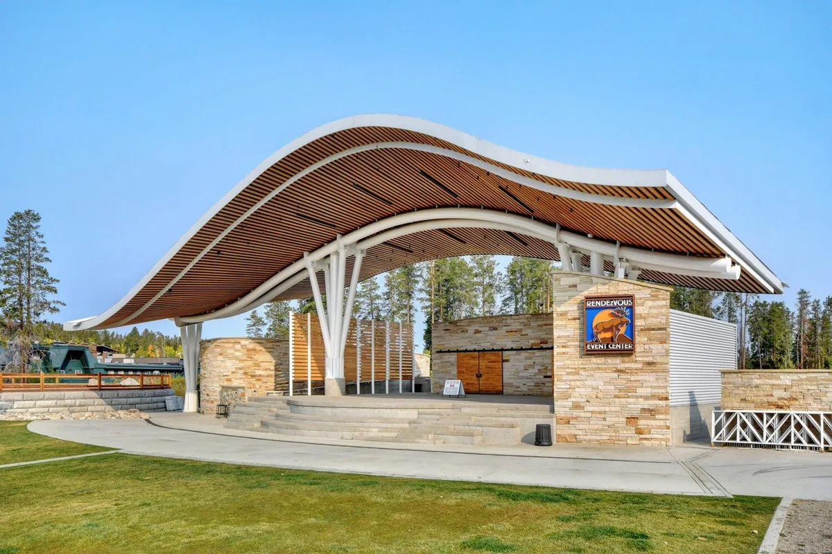 An exterior shot of the Hideaway Park amphitheater, featuring its distinctive curved roof and stone facade. Event signage hints at concerts or community gatherings, and the mood is festive and welcoming, representing local art showcases and outdoor celebrations.
